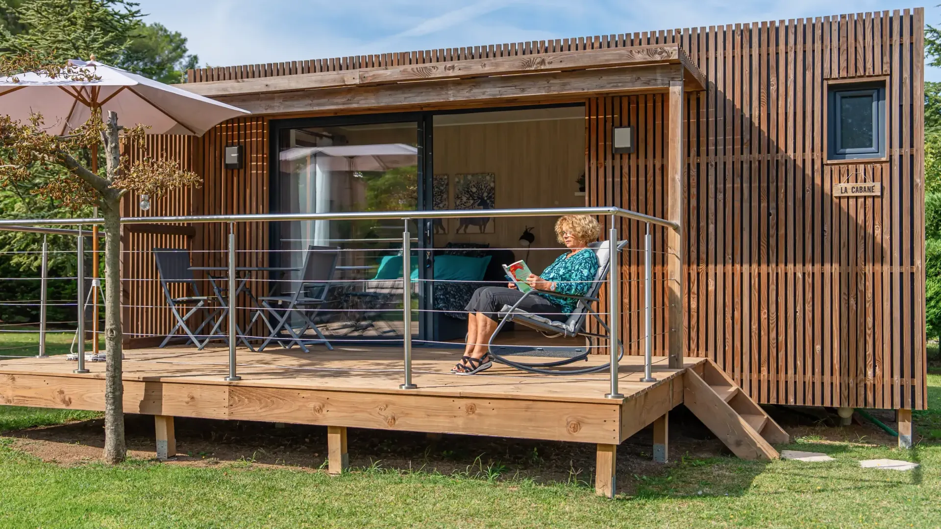 Studio de jardin Greenkub en ossature bois aménagé en chambre d'amis : une invitée lit sur la terrasse, baie coulissante ouverte