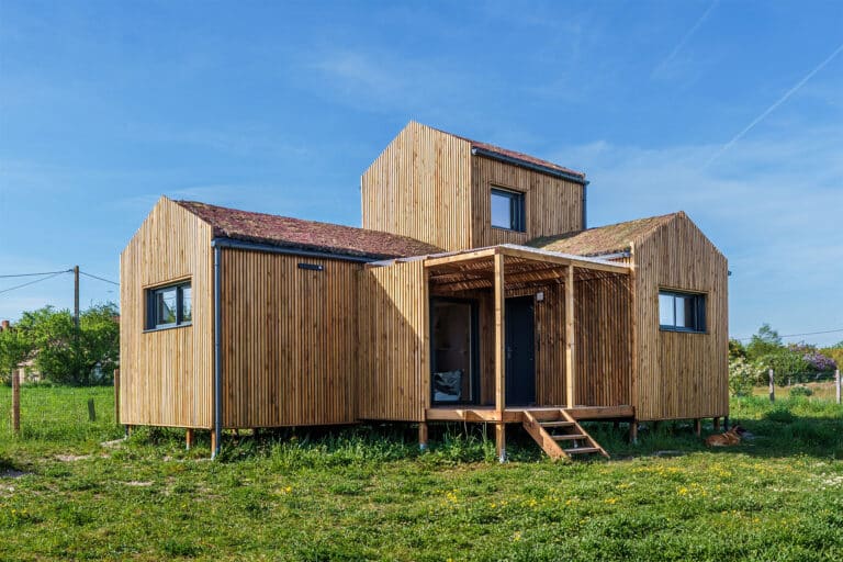 Façade principale d'une maison modulaire bois Greenkub GK50 – bardage bois vertical, toiture végétalisée multi-volumes, terrasse couverte avec pergola, structure sur plots métalliques