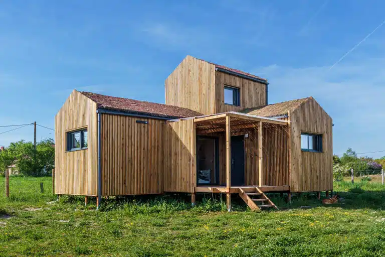 Façade principale d'une maison modulaire bois Greenkub GK50 – bardage bois vertical, toiture végétalisée multi-volumes, terrasse couverte avec pergola, structure sur plots métalliques