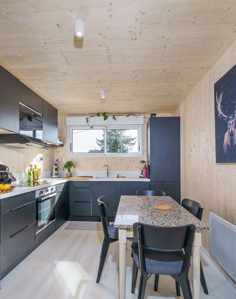 Cuisine équipée anthracite d'une maison modulaire bois Greenkub GK50 – murs CLT épicéa, plan de travail blanc, table de repas avec plateau granit, spots plafond cylindriques blancs