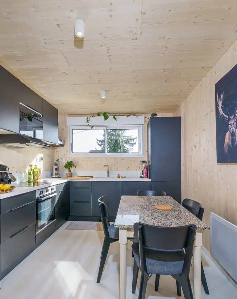 Cuisine équipée anthracite d'une maison modulaire bois Greenkub GK50 – murs CLT épicéa, plan de travail blanc, table de repas avec plateau granit, spots plafond cylindriques blancs