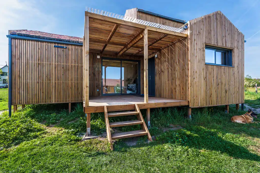 Façade principale d'une maison modulaire bois Greenkub GK50 – bardage bois vertical, toiture végétalisée multi-volumes, terrasse couverte avec pergola, structure sur plots métalliques
