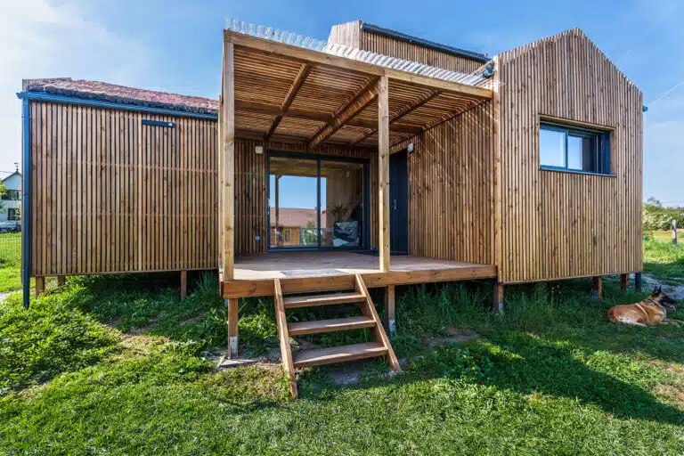 Façade principale d'une maison modulaire bois Greenkub GK50 – bardage bois vertical, toiture végétalisée multi-volumes, terrasse couverte avec pergola, structure sur plots métalliques