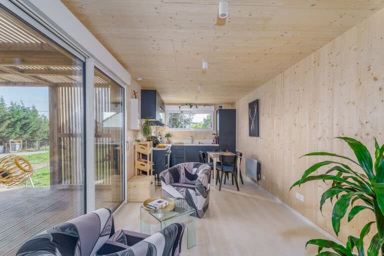 Intérieur salon-cuisine d'une maison modulaire bois Greenkub GK50 – murs et plafond en CLT épicéa naturel, cuisine en L anthracite, baie vitrée coulissante ouvrant sur terrasse bois