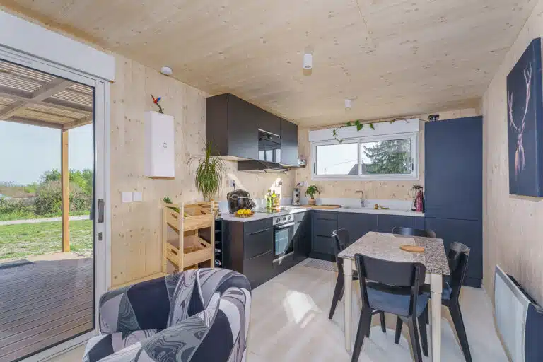 Intérieur maison bois CLT Greenkub – séjour lumineux avec baie vitrée, cuisine équipée noire et murs bois naturel